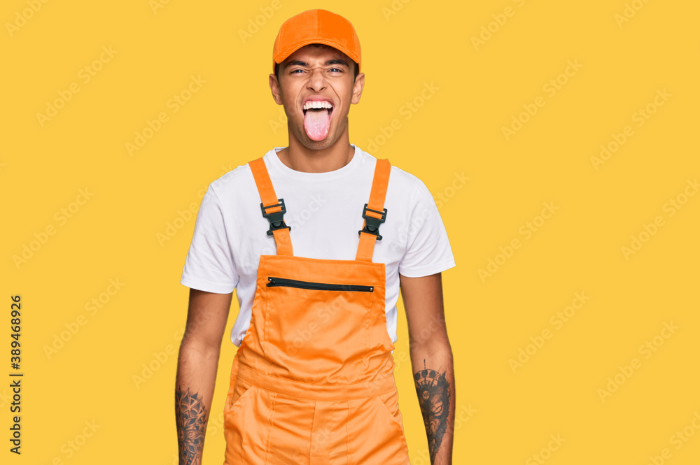 Young handsome african american man wearing handyman uniform sticking tongue out happy with funny expression. emotion concept. Young handsome african american man wearing handyman uniform sticking tongue out happy with funny expression. emotion concept.