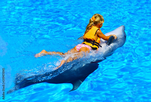 A little girl riding a dolphin rides in the oceanarium with blue and transparent water. Joy. Pleasure. Water splashes and bubbles. The child plays with a cute sea whale, holding tightly to the fins.