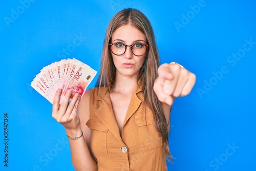 Obraz na plátně Young blonde girl holding israeli shekels pointing with finger to the camera and