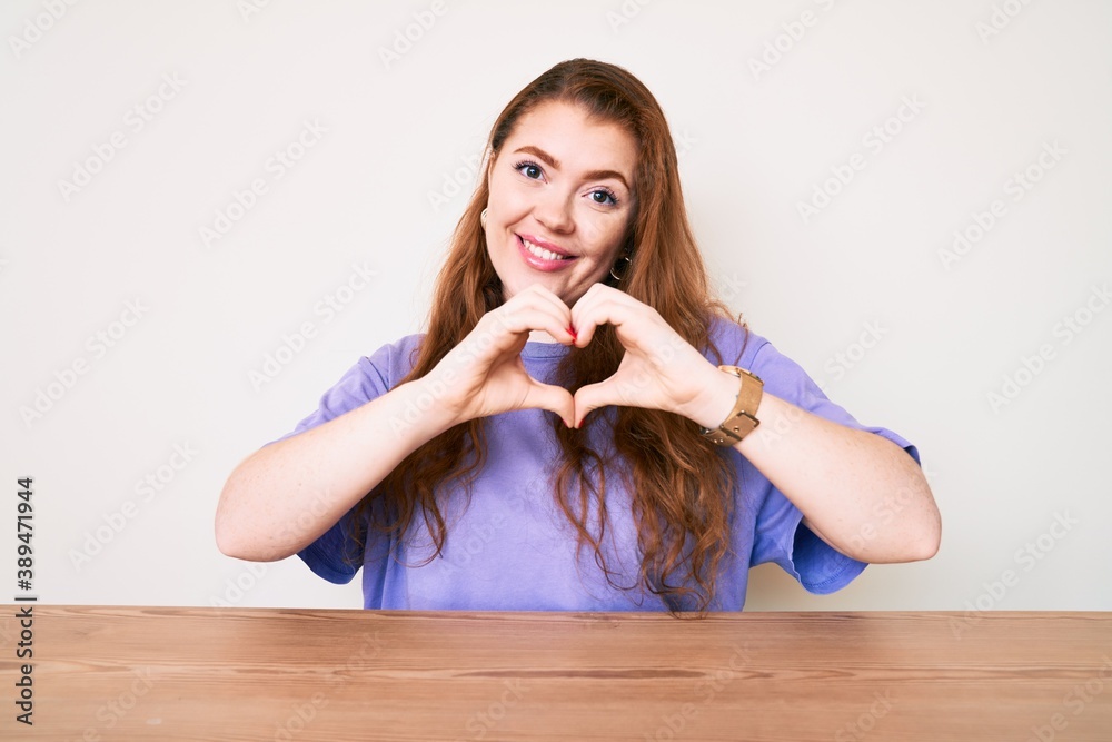 Young redhead woman wearing casual clothes sitting on the table smiling in love showing heart symbol and shape with hands. romantic concept. Young redhead woman wearing casual clothes sitting on the table smiling in love showing heart symbol and shape with hands. romantic concept.