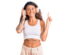 Young hispanic woman with tattoo listening to music using headphones surprised with an idea or question pointing finger with happy face, number one