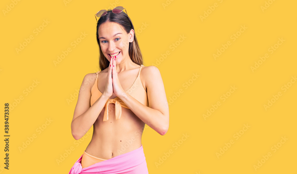 Fototapeta premium Beautiful brunette young woman wearing bikini and sunglasses praying with hands together asking for forgiveness smiling confident.