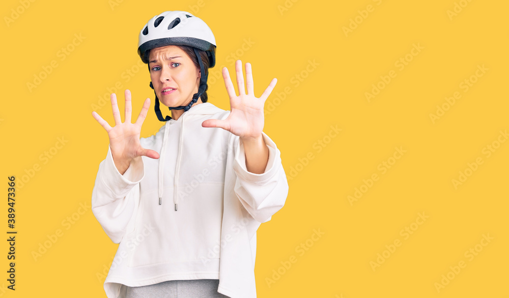 Beautiful brunette young woman wearing bike helmet and sporty clothes afraid and terrified with fear expression stop gesture with hands, shouting in shock. panic concept.