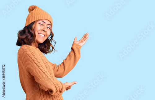 Young beautiful mixed race woman wearing wool sweater and winter hat inviting to enter smiling natural with open hand