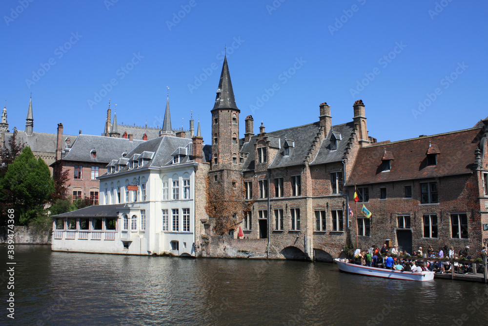 River side view of the old historic buildings of Bruges in Belgium on a sunny day. There is a boat with tourists on the river.