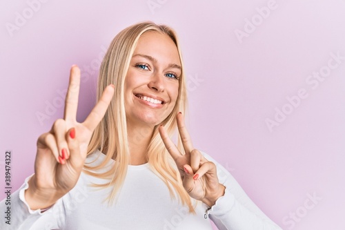 Young blonde girl wearing casual clothes smiling looking to the camera showing fingers doing victory sign. number two.