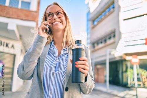 Young blonde businesswoman talking on the smartphone and holding bottle of water at the city.
