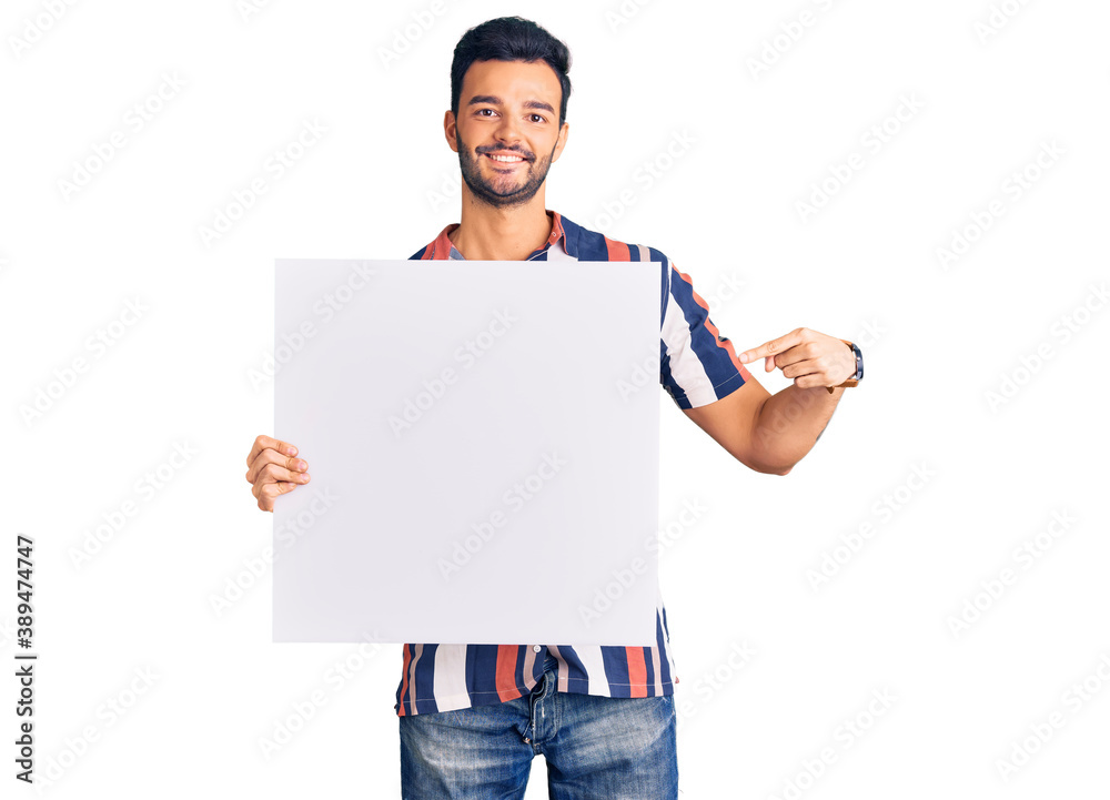 Young handsome hispanic man holding cardboard banner with blank space smiling happy pointing with hand and finger Young handsome hispanic man holding cardboard banner with blank space smiling happy pointing with hand and finger