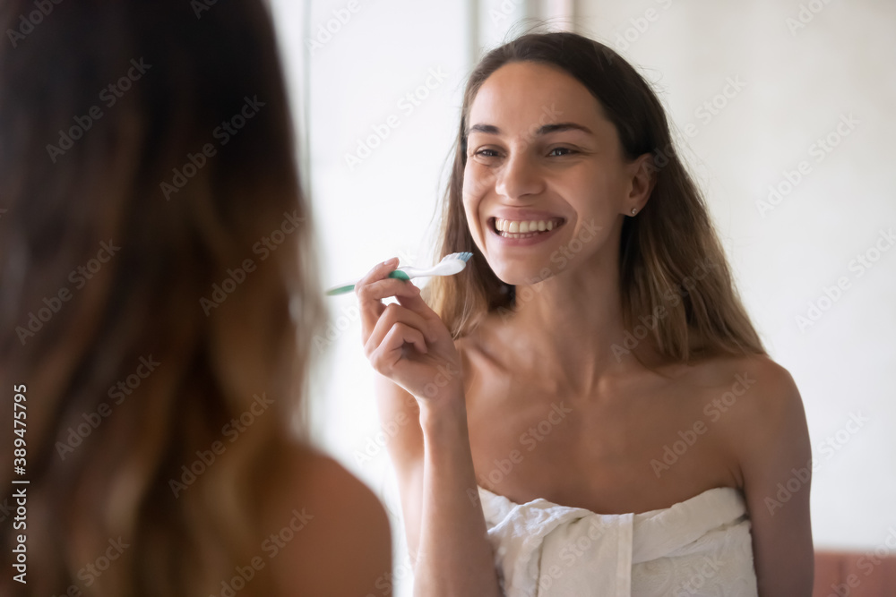 Healthy teeth. Pretty young millennial female standing in bathroom after taking shower and cleaning teeth at morning, looking at wide wall mirror with shiny beautiful smile holding toothbrush in hand Healthy teeth. Pretty young millennial female standing in bathroom after taking shower and cleaning teeth at morning, looking at wide wall mirror with shiny beautiful smile holding toothbrush in hand