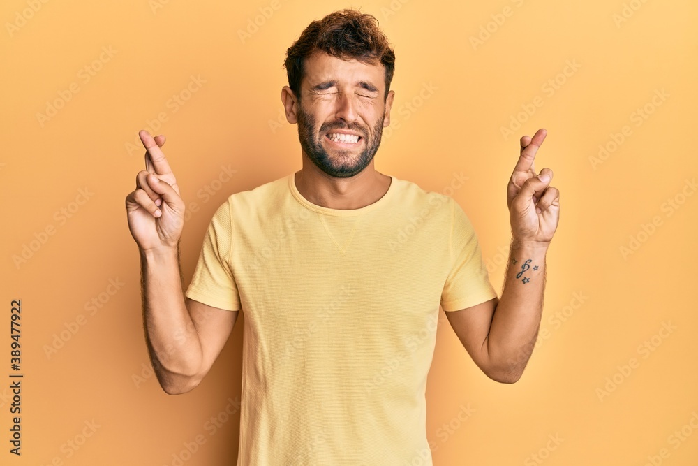 Handsome man with beard wearing casual yellow tshirt over yellow background gesturing finger crossed smiling with hope and eyes closed. luck and superstitious concept. Handsome man with beard wearing casual yellow tshirt over yellow background gesturing finger crossed smiling with hope and eyes closed. luck and superstitious concept.