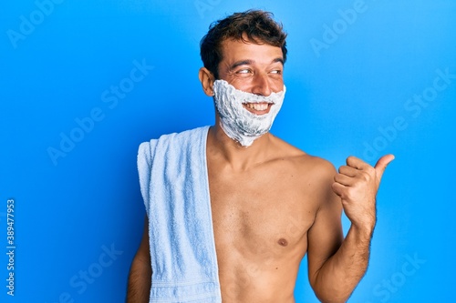 Handsome man saving beard with shave foam over face smiling with happy face looking and pointing to the side with thumb up.