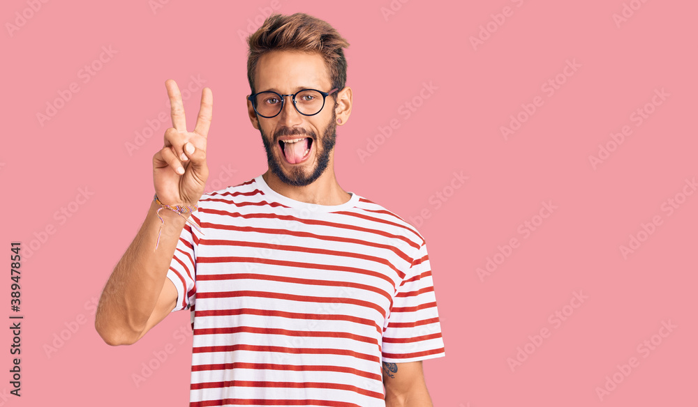 Naklejka premium Handsome blond man with beard wearing casual clothes and glasses smiling with happy face winking at the camera doing victory sign with fingers. number two.