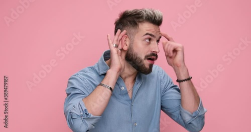 young casual man leaning to hear better but feeling disappointed of the crazy things he heard on pink background