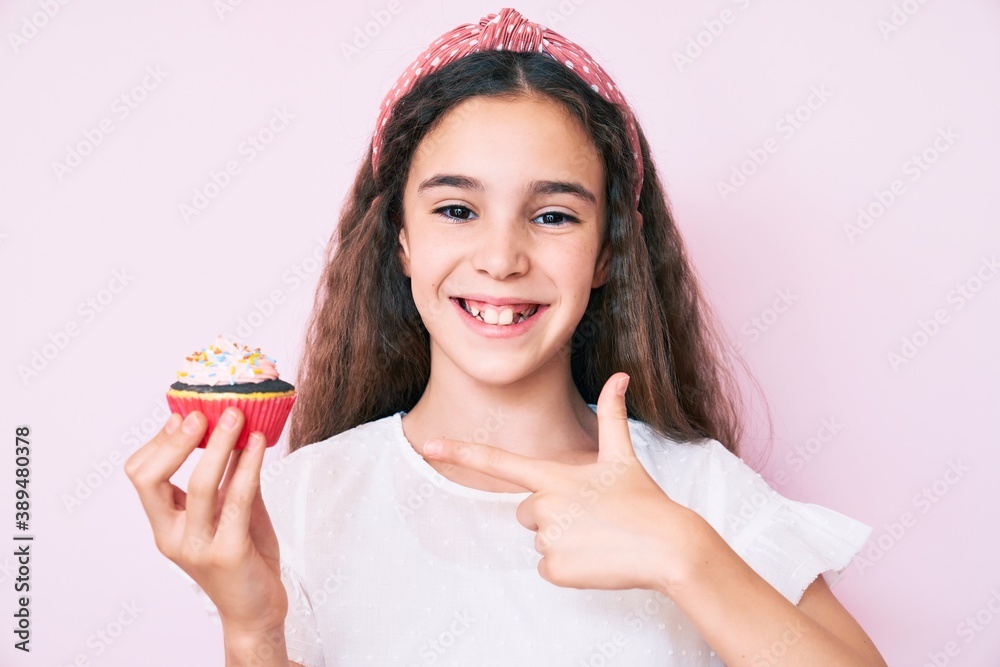 Cute hispanic child girl holding cupcake smiling happy pointing with hand and finger Cute hispanic child girl holding cupcake smiling happy pointing with hand and finger