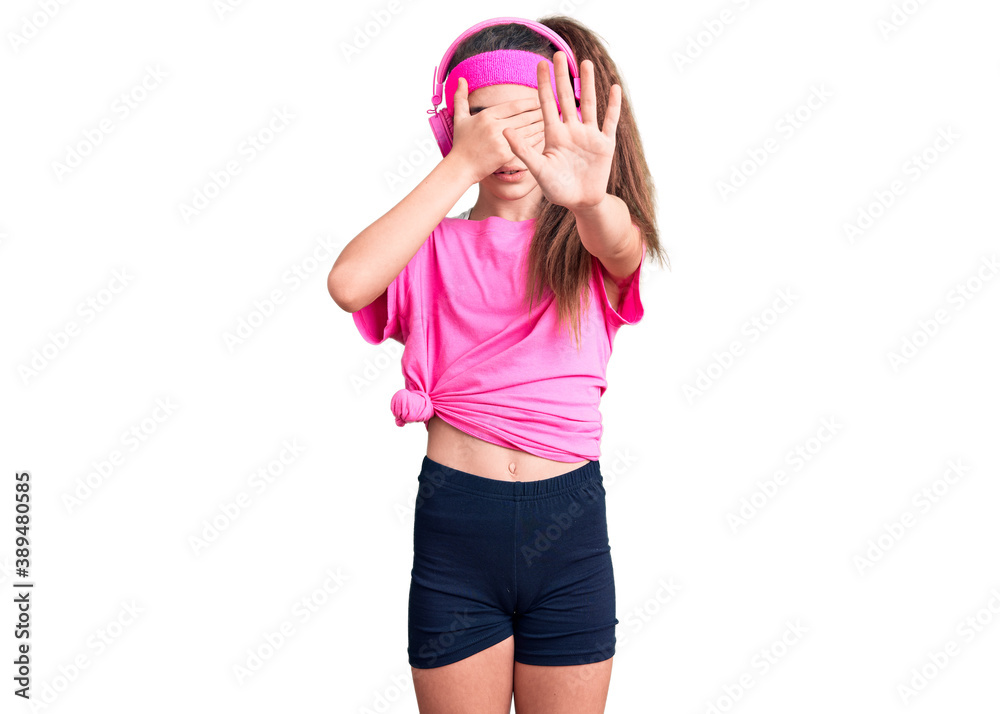 Cute hispanic child girl wearing gym clothes and using headphones covering eyes with hands and doing stop gesture with sad and fear expression. embarrassed and negative concept. Cute hispanic child girl wearing gym clothes and using headphones covering eyes with hands and doing stop gesture with sad and fear expression. embarrassed and negative concept.