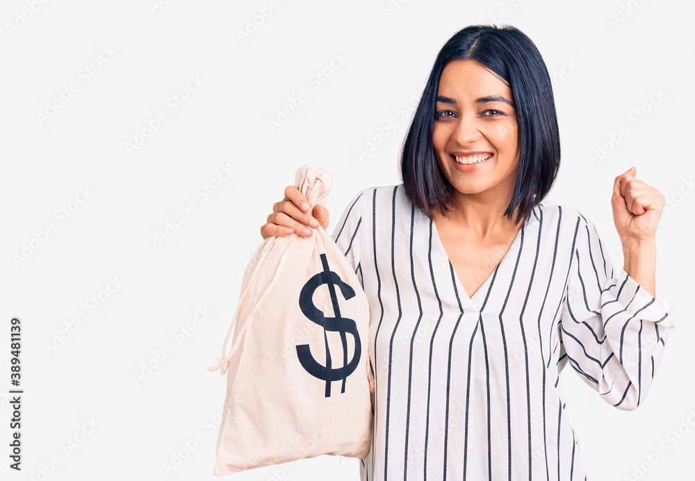 Young beautiful latin woman holding money bag with dollar symbol screaming proud, celebrating victory and success very excited with raised arms Young beautiful latin woman holding money bag with dollar symbol screaming proud, celebrating victory and success very excited with raised arms