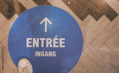 sign on a wooden background.

Forward arrow labeled in French and English: Ingang, in a blue circle with a white sneaker on a wood floor, space for text on the right, top view. photo