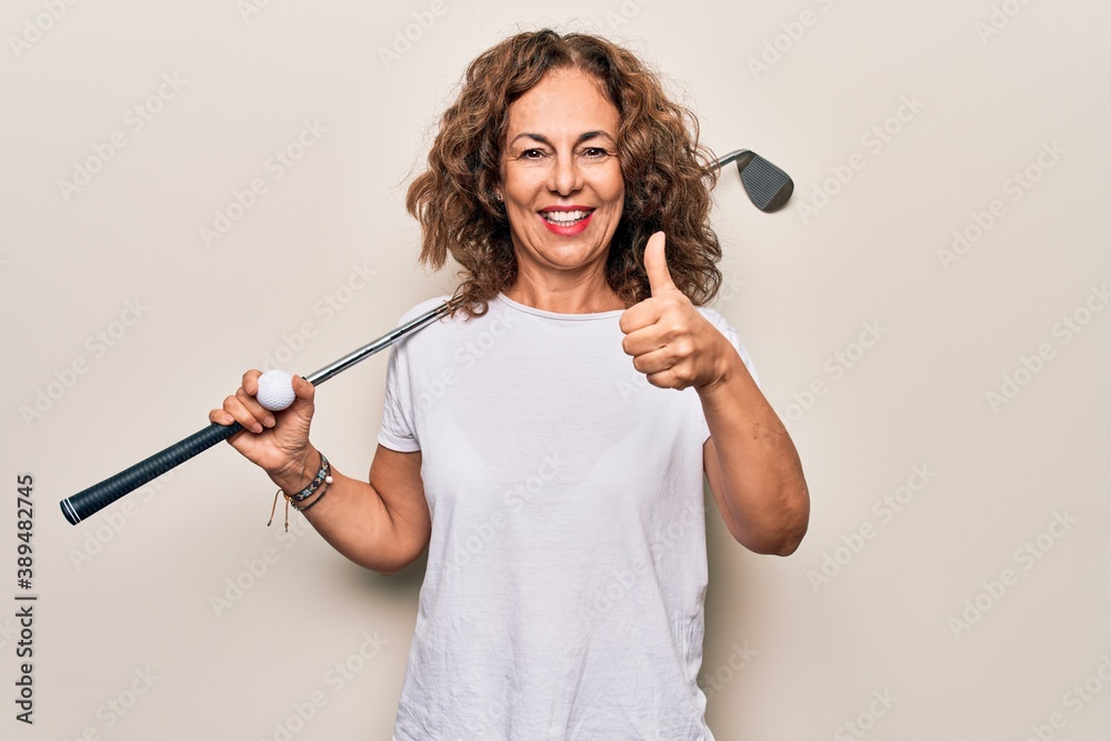 Middle age beautiful sportswoman playing golf using stick and ball over white background smiling happy and positive, thumb up doing excellent and approval sign Middle age beautiful sportswoman playing golf using stick and ball over white background smiling happy and positive, thumb up doing excellent and approval sign