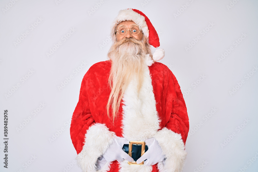 Old senior man with grey hair and long beard wearing santa claus costume with suspenders puffing cheeks with funny face. mouth inflated with air, crazy expression.