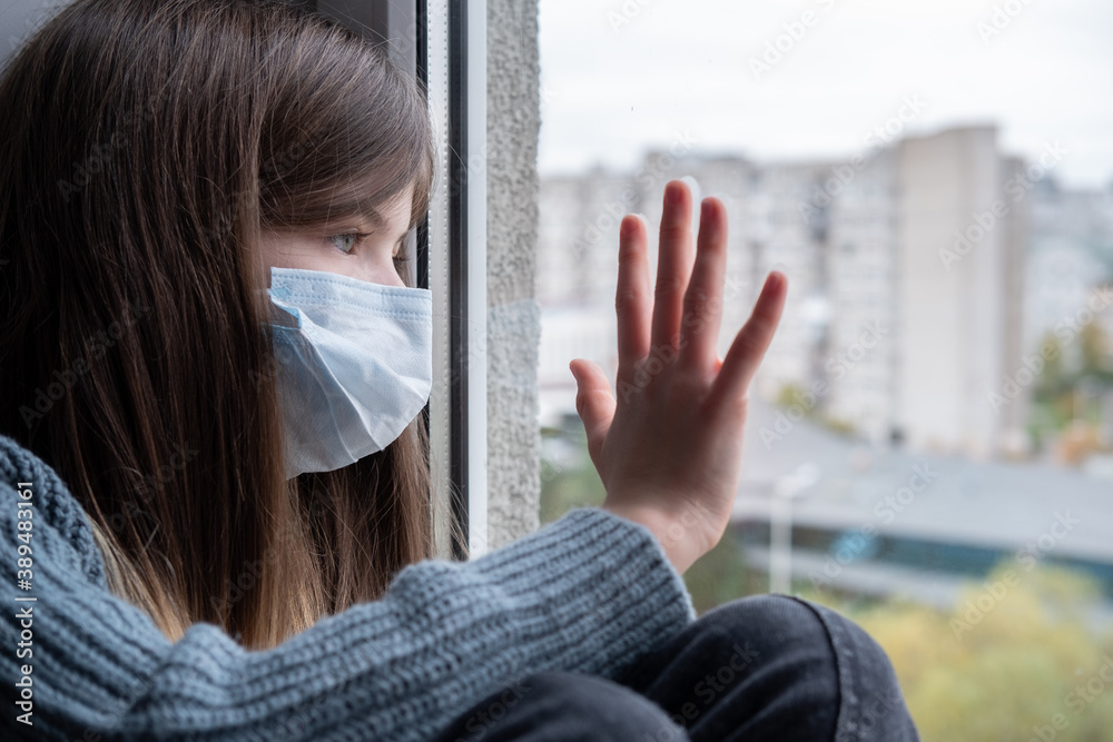 a little girl in a medical mask is sitting by the window, quarantined. World coronavirus pandemic, social isolation, quarantine concept