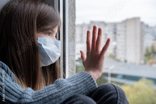 Obraz na płótnie a little girl in a medical mask is sitting by the window, quarantined. World coronavirus pandemic, social isolation, quarantine concept w salonie