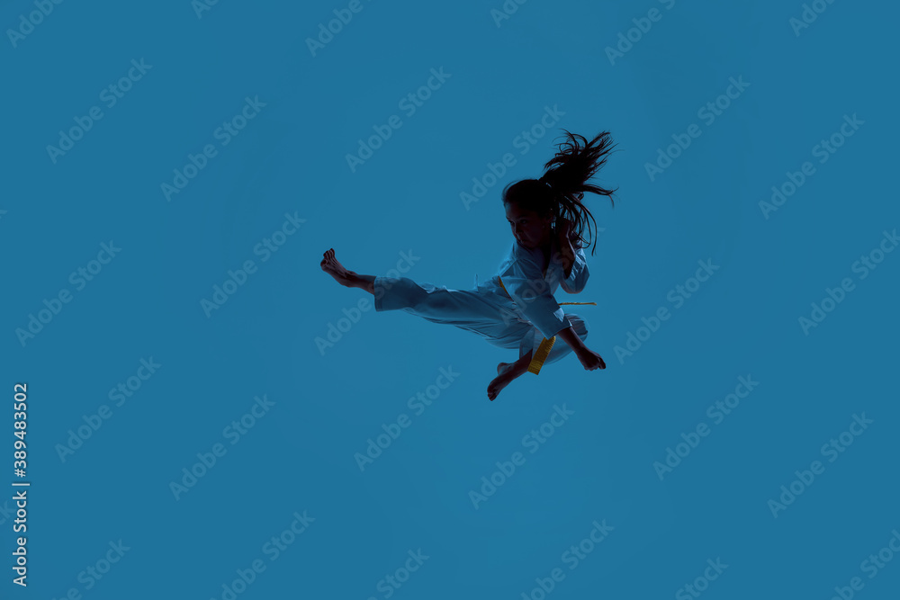 Silhouetted shot of little karate girl in white kimono exercising and fighting, doing martial arts, jumping isolated over blue background Silhouetted shot of little karate girl in white kimono exercising and fighting, doing martial arts, jumping isolated over blue background