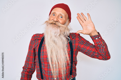 Old senior man with grey hair and long beard wearing hipster look with wool cap waiving saying hello happy and smiling, friendly welcome gesture