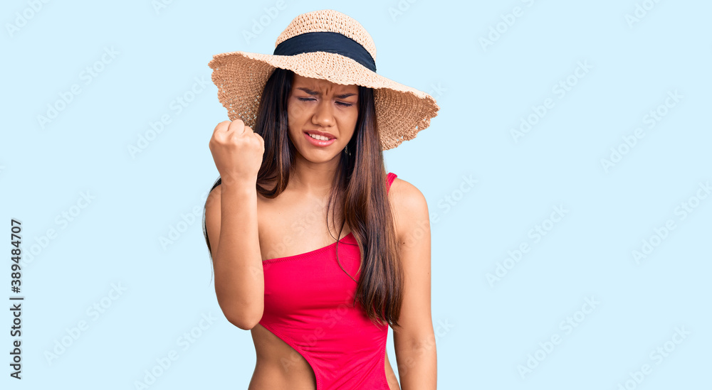Young beautiful latin girl wearing swimwear and summer hat angry and mad raising fist frustrated and furious while shouting with anger. rage and aggressive concept. Young beautiful latin girl wearing swimwear and summer hat angry and mad raising fist frustrated and furious while shouting with anger. rage and aggressive concept.
