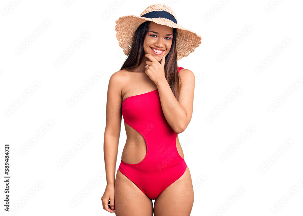 Young beautiful latin girl wearing swimwear and summer hat looking confident at the camera smiling with crossed arms and hand raised on chin. thinking positive. Young beautiful latin girl wearing swimwear and summer hat looking confident at the camera smiling with crossed arms and hand raised on chin. thinking positive.