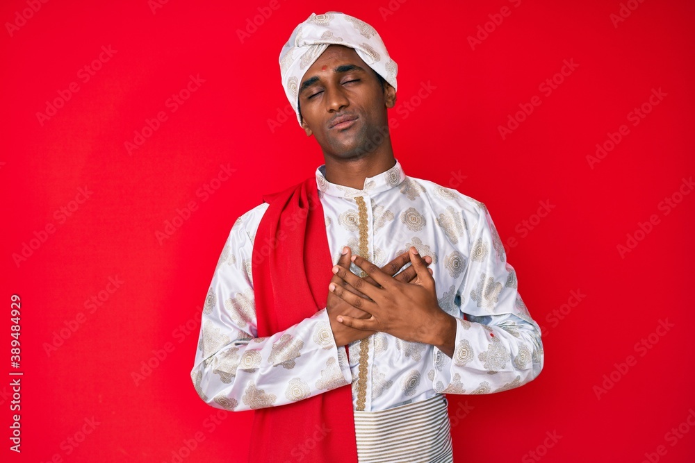 Handsome indian man wearing tradition sherwani saree clothes smiling with hands on chest with closed eyes and grateful gesture on face. health concept. Handsome indian man wearing tradition sherwani saree clothes smiling with hands on chest with closed eyes and grateful gesture on face. health concept.