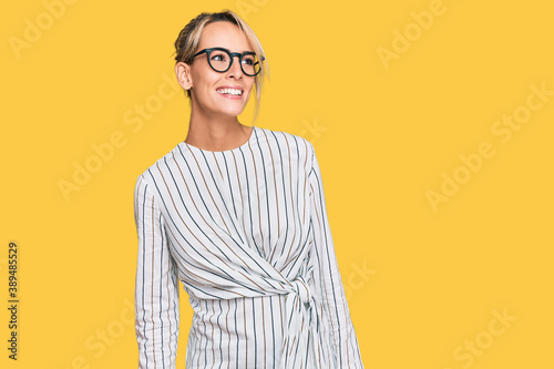 Beautiful blonde woman wearing business shirt and glasses looking away to side with smile on face, natural expression. laughing confident.