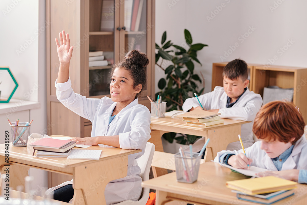Mixed-race girl of elementary age in whitecoat raising hand to answer question Mixed-race girl of elementary age in whitecoat raising hand to answer question