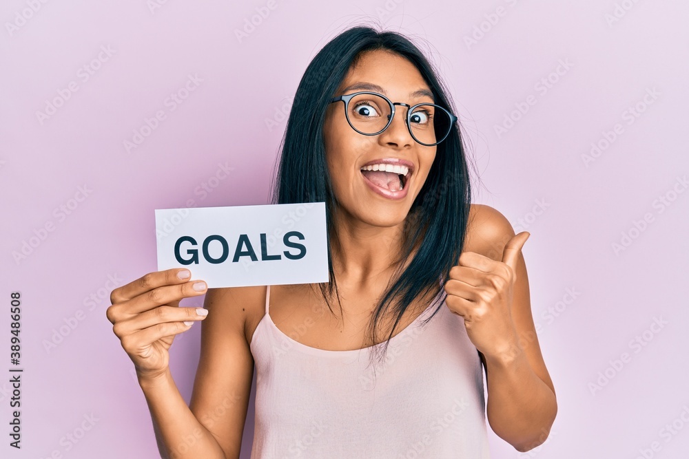 Young african american woman holding goals message paper pointing thumb up to the side smiling happy with open mouth Young african american woman holding goals message paper pointing thumb up to the side smiling happy with open mouth