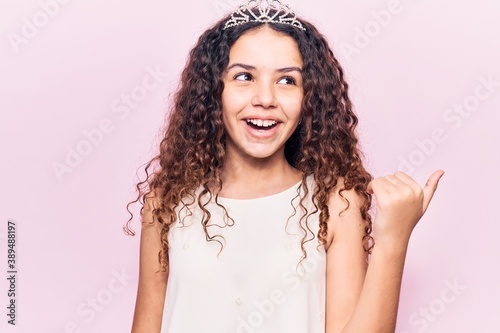 Beautiful kid girl with curly hair wearing princess tiara pointing thumb up to the side smiling happy with open mouth