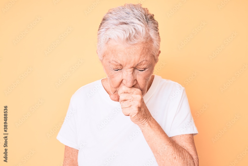 Senior beautiful woman with blue eyes and grey hair wearing classic white tshirt over yellow background feeling unwell and coughing as symptom for cold or bronchitis. health care concept. Senior beautiful woman with blue eyes and grey hair wearing classic white tshirt over yellow background feeling unwell and coughing as symptom for cold or bronchitis. health care concept.