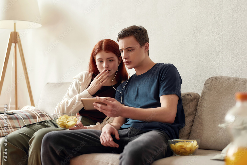 Couple of teenagers sitting on the couch at home, looking emotional while watching something on the phone, using the same pair of earphones