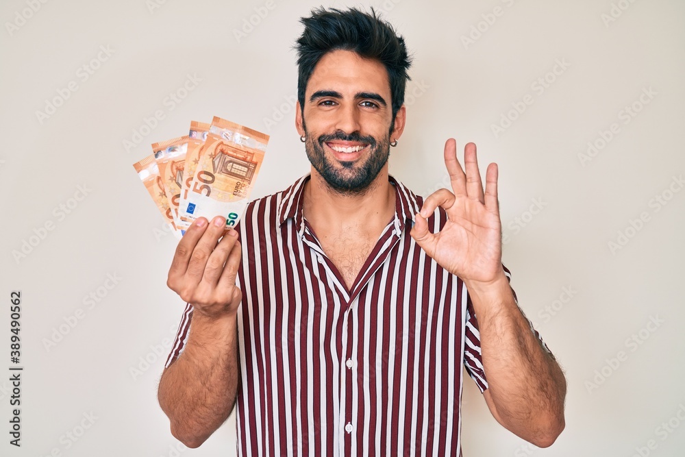 Handsome hispanic man with beard holding euro banknotes doing ok sign with fingers, smiling friendly gesturing excellent symbol Handsome hispanic man with beard holding euro banknotes doing ok sign with fingers, smiling friendly gesturing excellent symbol