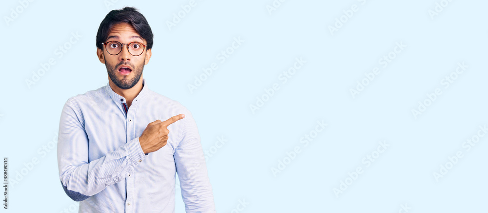 Handsome hispanic man wearing business shirt and glasses surprised pointing with finger to the side, open mouth amazed expression. Handsome hispanic man wearing business shirt and glasses surprised pointing with finger to the side, open mouth amazed expression.