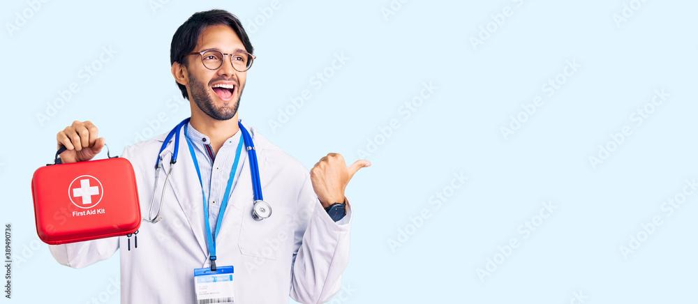 Handsome hispanic man wearing doctor coat holding first aid kit pointing thumb up to the side smiling happy with open mouth Handsome hispanic man wearing doctor coat holding first aid kit pointing thumb up to the side smiling happy with open mouth