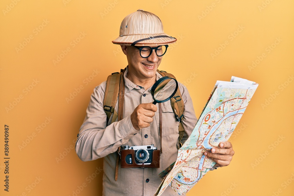 Middle age bald man wearing explorer hat holding magnifying glass on a map smiling with a happy and cool smile on face. showing teeth. Middle age bald man wearing explorer hat holding magnifying glass on a map smiling with a happy and cool smile on face. showing teeth.
