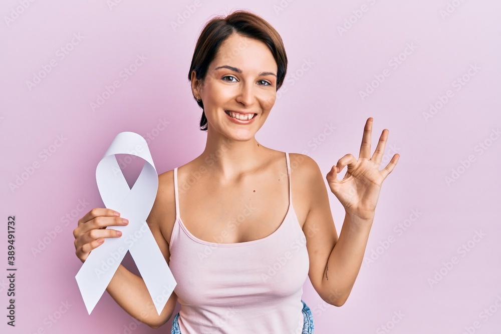 Young brunette woman with short hair holding cancer awareness white ribbon doing ok sign with fingers, smiling friendly gesturing excellent symbol Young brunette woman with short hair holding cancer awareness white ribbon doing ok sign with fingers, smiling friendly gesturing excellent symbol