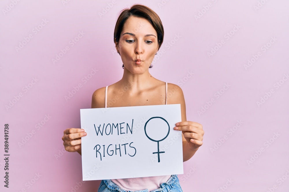 Young brunette woman with short hair holding woman rights banner making fish face with mouth and squinting eyes, crazy and comical. Young brunette woman with short hair holding woman rights banner making fish face with mouth and squinting eyes, crazy and comical.