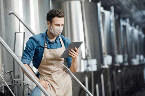 Busy millennial attractive man in apron and protective mask uses tablet to control brewery with mobile app