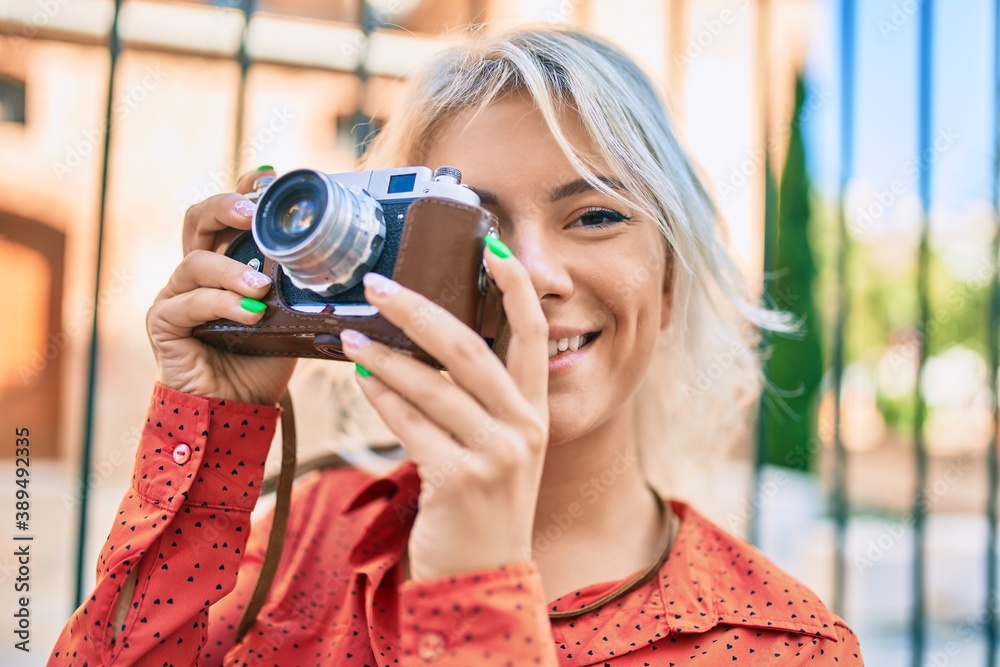Young blonde woman smiling happy using vintage camera walking at the city.