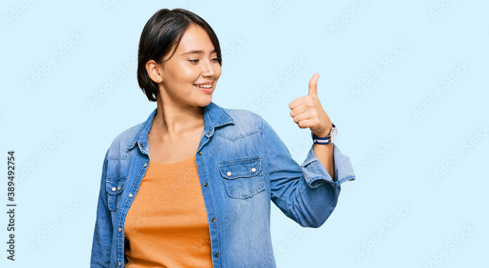 Young beautiful hispanic woman with short hair wearing casual denim jacket looking proud, smiling doing thumbs up gesture to the side Young beautiful hispanic woman with short hair wearing casual denim jacket looking proud, smiling doing thumbs up gesture to the side