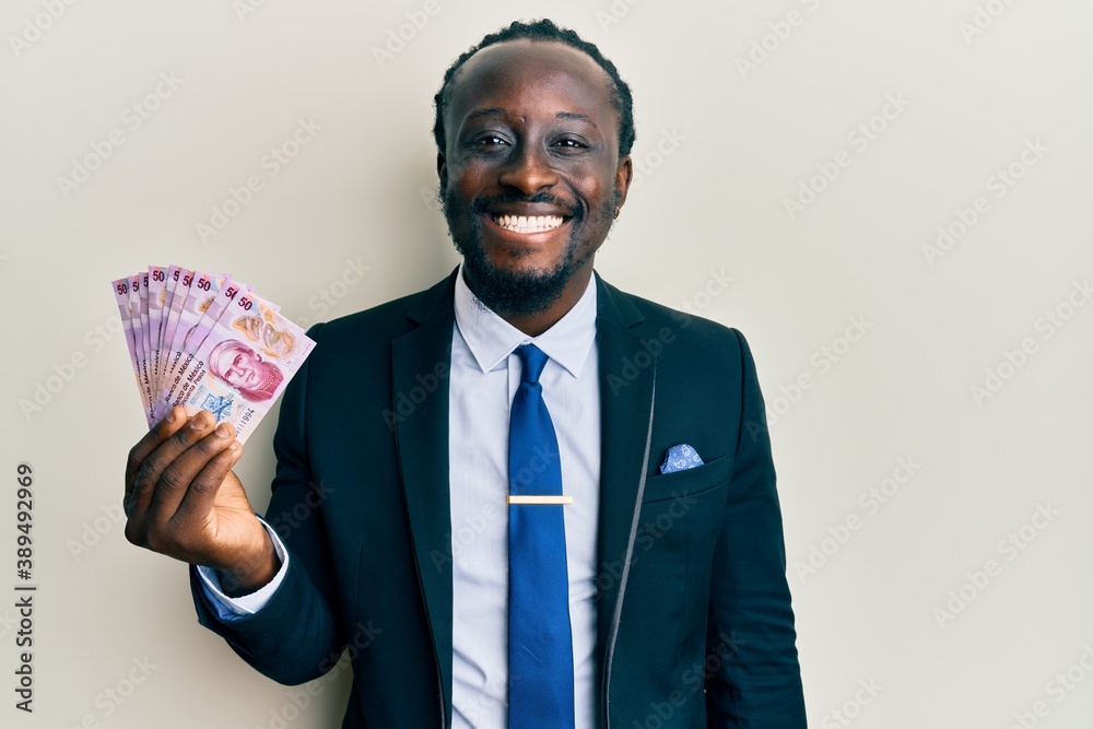 Handsome young black man wearing business suit holding mexican pesos banknotes looking positive and happy standing and smiling with a confident smile showing teeth Handsome young black man wearing business suit holding mexican pesos banknotes looking positive and happy standing and smiling with a confident smile showing teeth