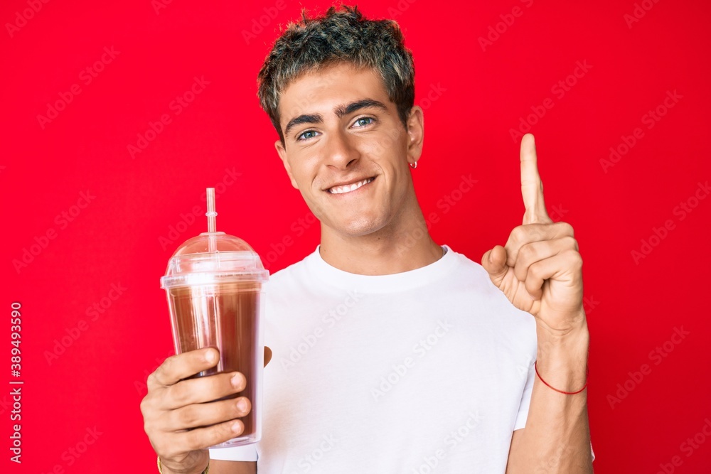 Young handsome man holding glass of smoothie smiling with an idea or question pointing finger with happy face, number one Young handsome man holding glass of smoothie smiling with an idea or question pointing finger with happy face, number one
