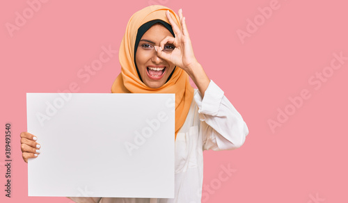 Young brunette arab woman wearing traditional islamic hijab holding banner smiling happy doing ok sign with hand on eye looking through fingers