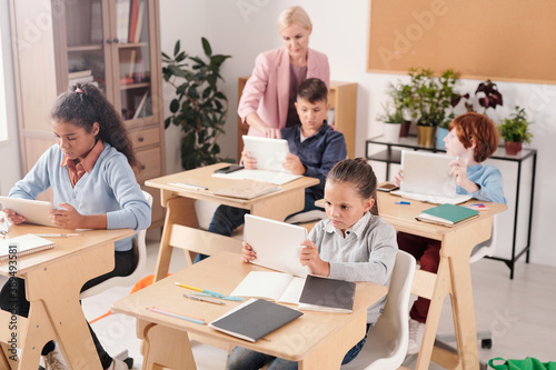 Serious schoolgirl and her classmates with tablets sitting by desks in classroom