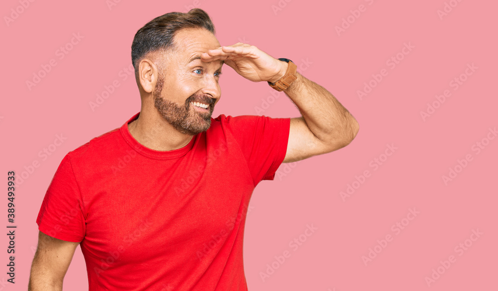 Handsome middle age man wearing casual red tshirt very happy and smiling looking far away with hand over head. searching concept.